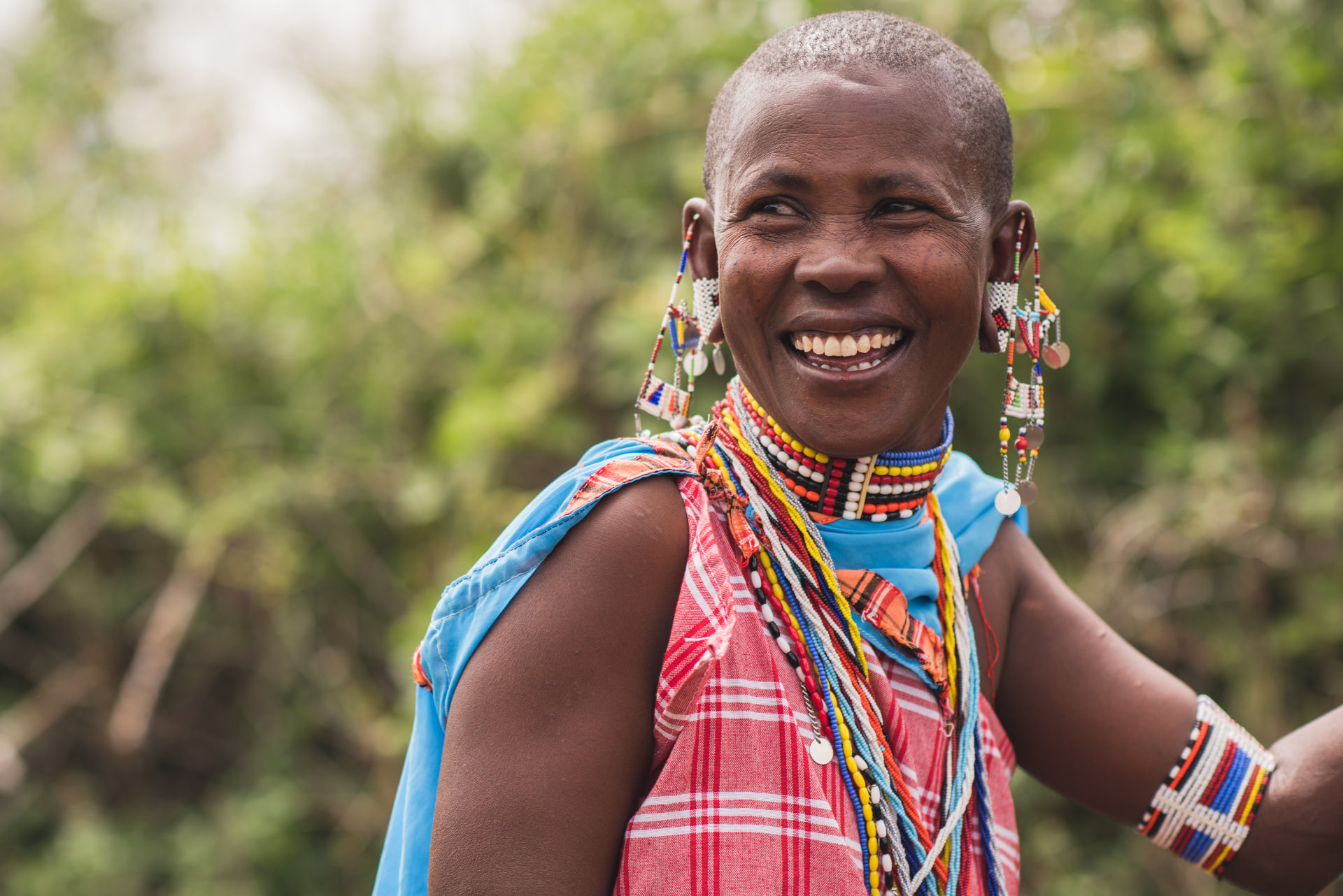 A Maasai woman wearing beaded necklaces, a beaded collar and hanging earrings smiles outdoors
